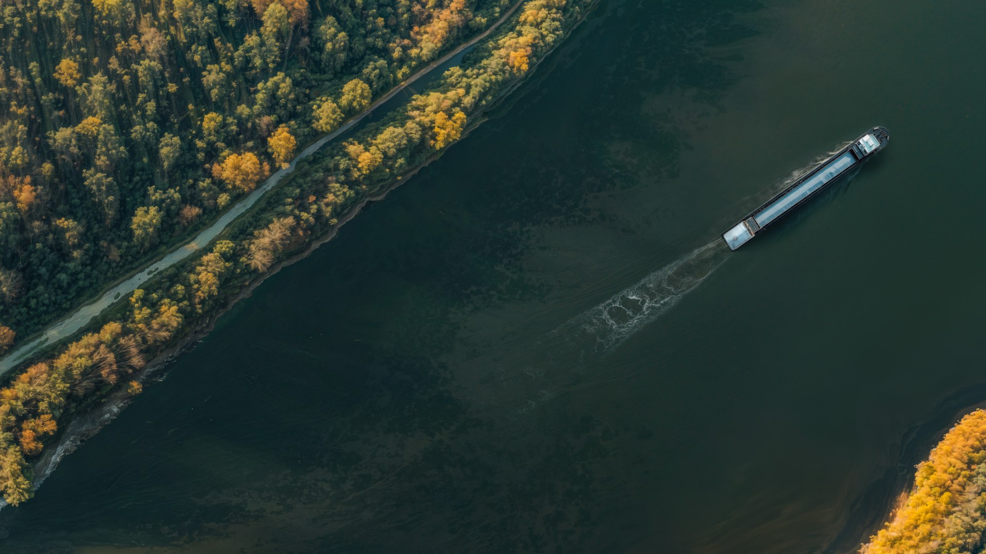 Aerial view of a cargo vessel traveling along a river bordered by forested shoreline