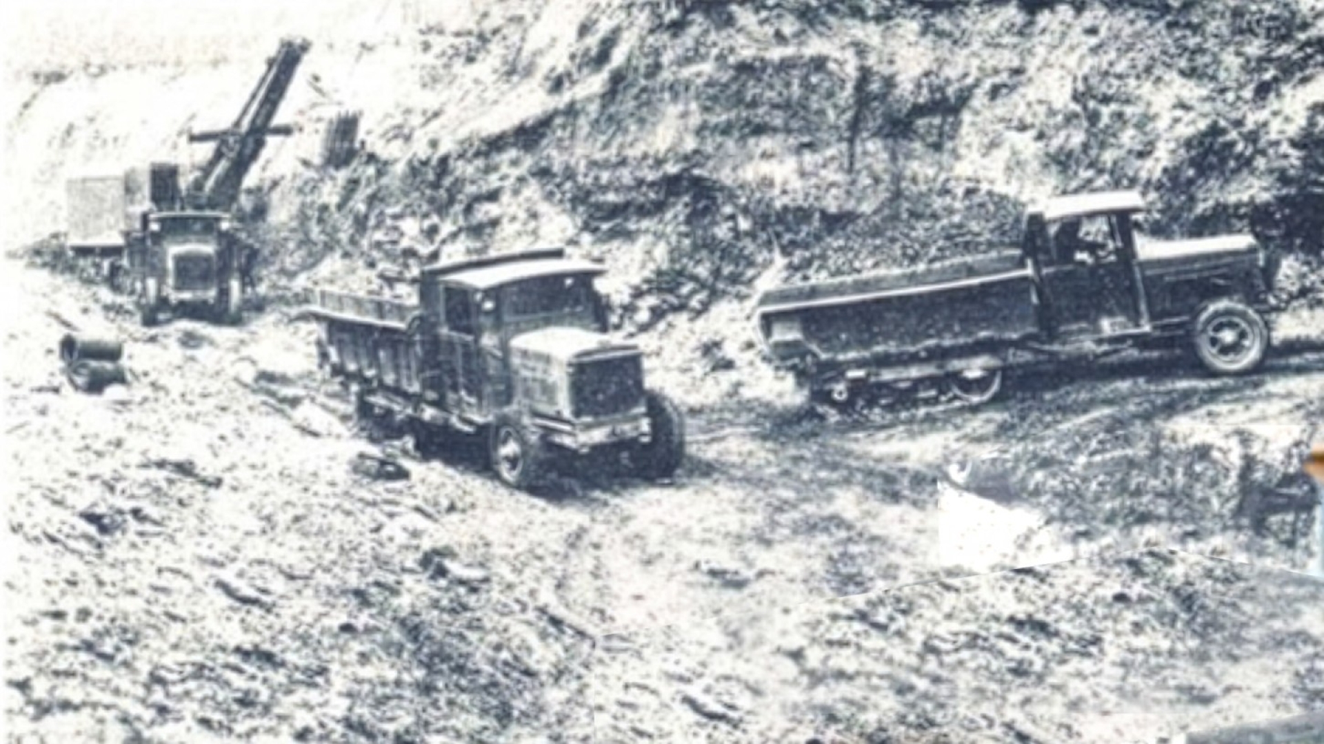 Vintage dump trucks and an excavator operating on a muddy construction road cut into a hillside