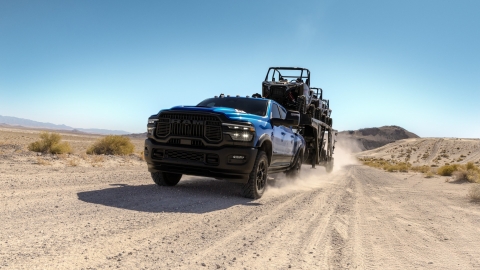 Blue Ram pickup truck towing a trailer carrying off-road vehicles on a dusty desert road
