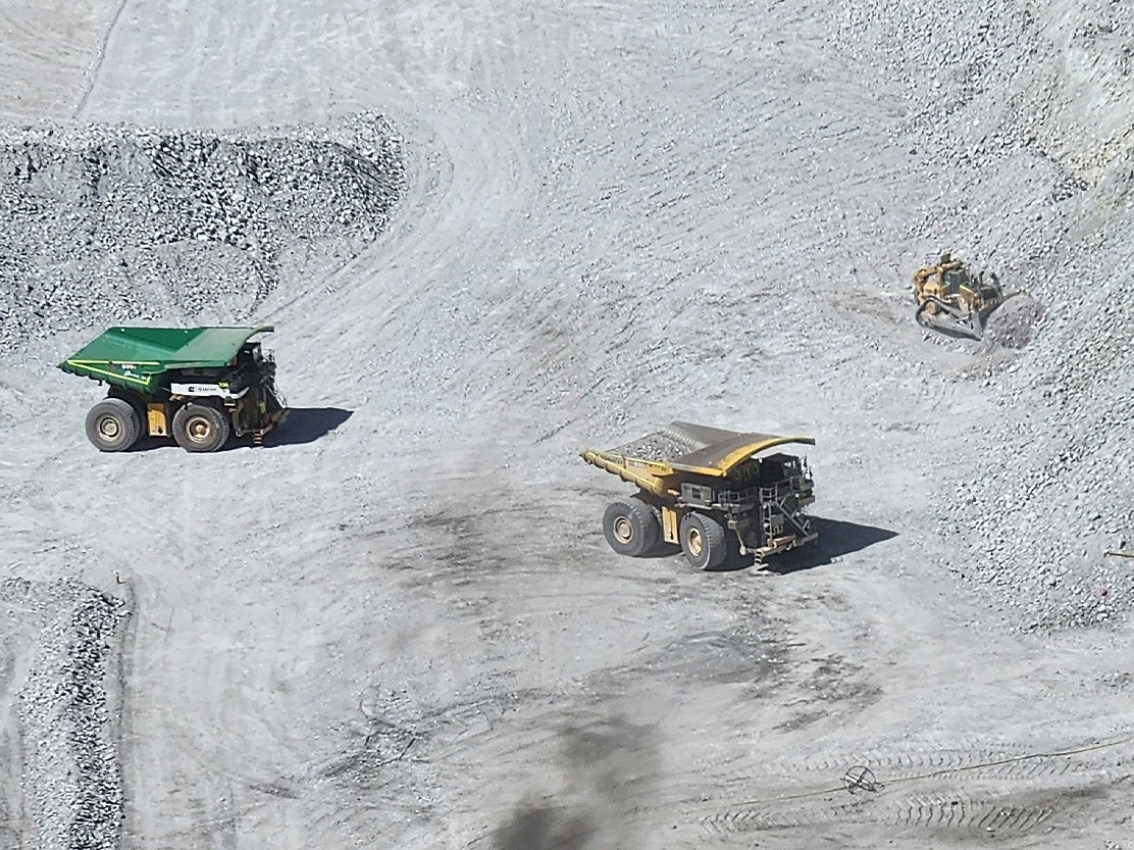 Aerial view of two large mining haul trucks and a bulldozer operating on a light-colored open-pit mine floor