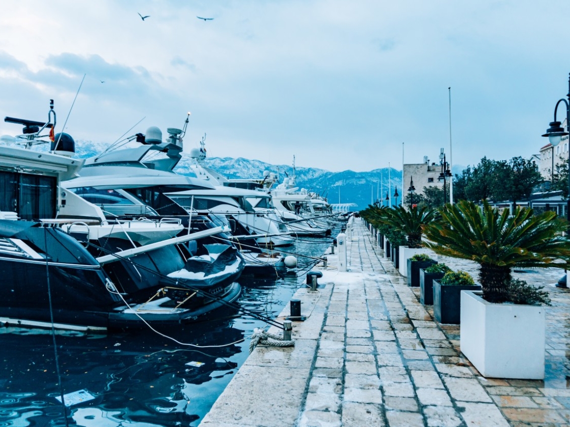 Yachts docked at a marina beside a waterfront walkway with mountains in the distance